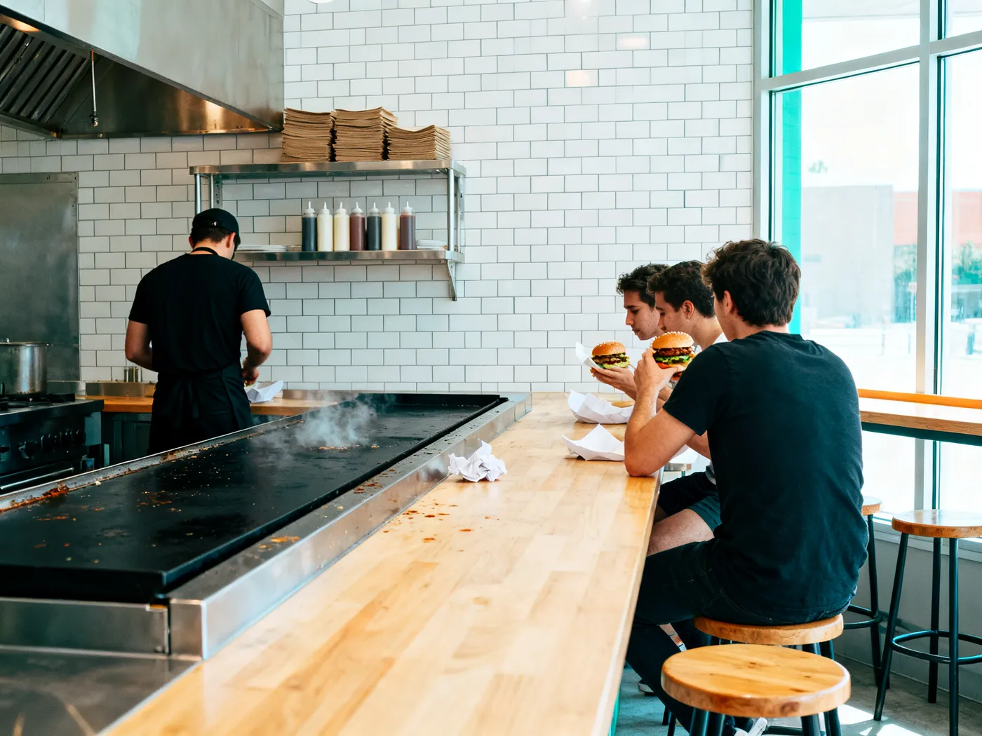 Inside in daylight: a long pale wood counter with stools, customers eating, and a cook at the flat-top griddle against white tiles.